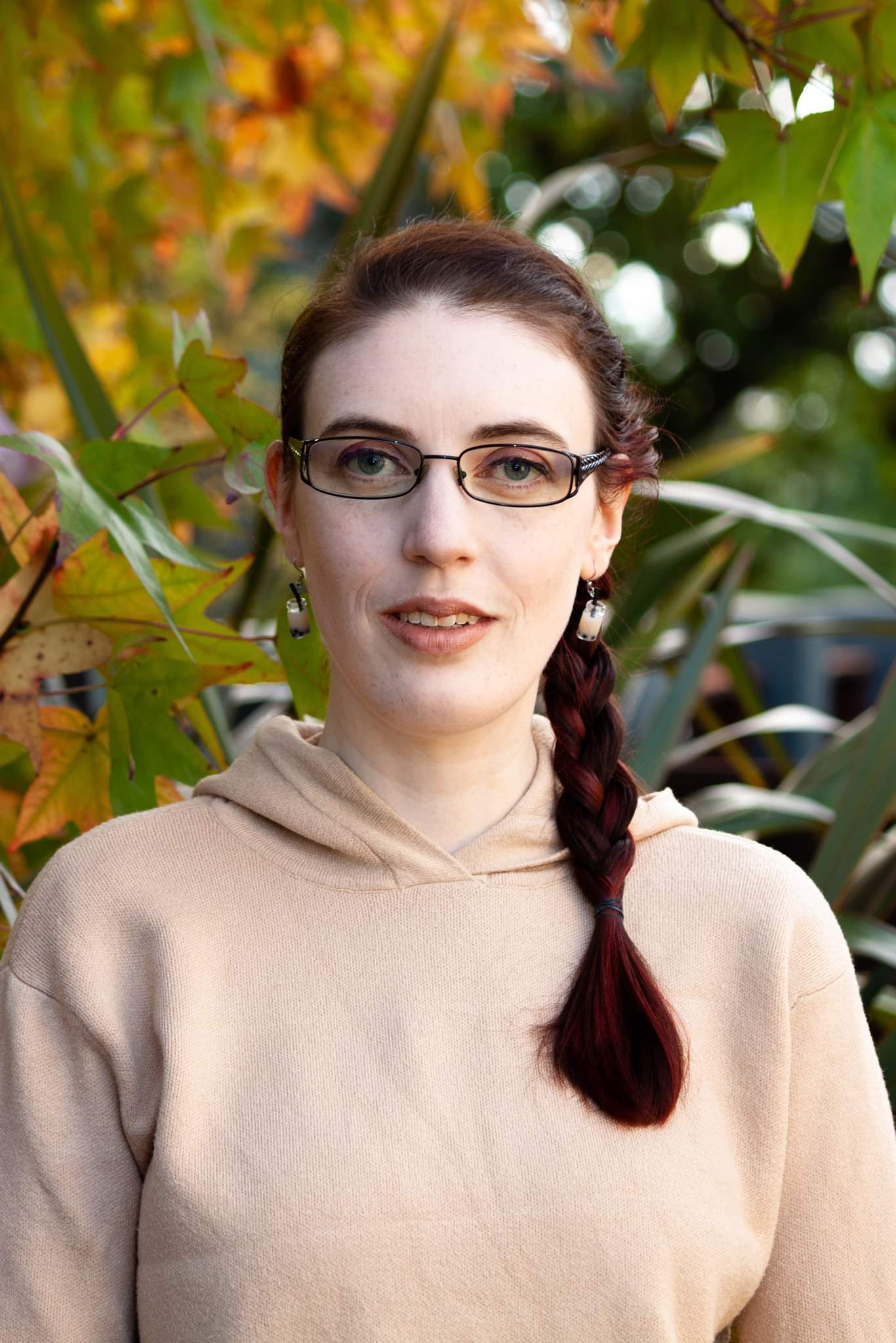 A photo of a pale person with dark brown and red hair in a braid. She has blue eyes and wears glasses. Her beige knit hoodie matches the bubble tea earrings she is wearing. There is yellow and green foliage in the background behind her. A photo of a pale person with glasses